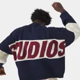 Sweater de punto grueso color azul marino con franja horizontal en el pecho que contiene la palabra "EME" en color bordó, bordeada en blanco. Las mangas son de color azul marino con una franja ancha color crema/blanco roto en la parte superior del brazo.