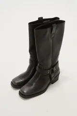 Bota de caña media color negro, estilo motociclista o cowboy, con puntera cuadrada y taco bajo. Presenta una tira ancha con hebillas metálicas alrededor del tobillo.