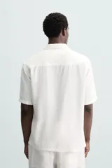 Camisa de calce relajado confeccionada en tejido fluido de mezcla de viscosa, color blanco. Presenta cuello bowling, manga corta, aberturas laterales en el bajo y cierre frontal con botones al tono.