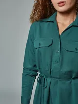 Vestido camisero verde con cuello clásico, mangas largas, cierre frontal con botones, dos bolsillos con solapa en el pecho y cinturón con lazo.