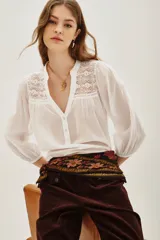 Camisa blanca de algodón con cuello mao, escote en V y cierre de botones. Presenta detalles de encaje calado en los hombros y mangas largas abullonadas con puños elásticos.