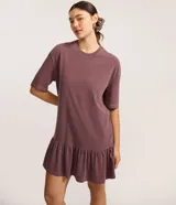 Vestido tipo camiseta de algodón, color malva o ciruela con efecto lavado, de corte recto y holgado, con cuello redondo y mangas cortas. Presenta un volado fruncido en el bajo.