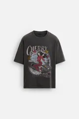Camiseta negra con cuello redondo y mangas cortas. Estampado frontal con la frase "Cherry Pill Blush Delight" y una ilustración de una mujer en un cohete. Tiene un efecto de lavado que le da una apariencia única.