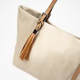 Bolso tipo shopper color beige con textura de espiga y detalles en cuero color camel. Tiene doble asa de hombro y un dije de borla en uno de los laterales.