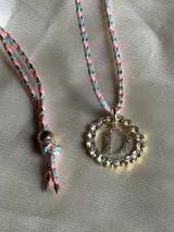 Collar con dije circular dorado con strass y letra 'A' en el centro, con cordón trenzado multicolor y cierre ajustable.