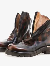 Bota corta de cuero bovino con efecto envejecido en tonos marrón oscuro y caramelo. Presenta cierre frontal con cremallera y suela de goma gruesa con relieve. El tacón bajo tiene un detalle metálico horizontal en color cobre.