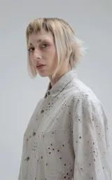 Camisa de manga corta color gris claro con cuello camisero y botones frontales. La tela presenta un bordado tipo broderie con motivos florales y geométricos.