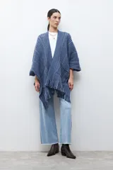 Ruana de tejido de punto en tonos beige y blanco, con patrón de espiga. Presenta cuello en V, mangas amplias, dos bolsillos frontales y flecos en el dobladillo.