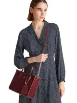 Cartera tipo tote bag de color borgoña, con acabado liso y detalles en cuero sintético del mismo tono. Presenta dos asas superiores y una correa ajustable extraíble. Incluye un colgante decorativo con el escudo de Hilfiger bordado y un dije metálico con el monograma TH.