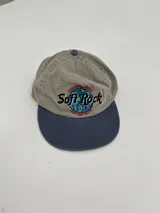 Gorro tipo gorra de béisbol, color gris claro con visera azul. Presenta un bordado frontal con el texto "Soft Rock Cafe" y un diseño de un globo terráqueo con detalles en rojo, azul y verde.