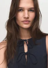 Vestido corto sin mangas de efecto denim, color azul oscuro, con cuello redondo y detalle de lazos ajustables en el escote frontal. Presenta un diseño con tablas que le dan volumen a la falda.