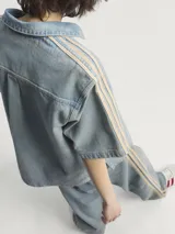 Camisa de manga corta Adidas Premium Essentials, confeccionada en denim azul claro 100% algodón. Diseño inspirado en la ropa de trabajo, con cuello clásico, cierre frontal con botones, bolsillo de parche en el pecho y logo bordado en el mismo tono. Presenta las tres tiras características de la marca en color beige en los hombros.