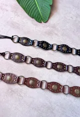Cinto de cuero con diseño occidental, compuesto por secciones ovaladas unidas por anillos metálicos. Cada sección presenta un medallón central con relieve y tachas metálicas alrededor. Disponible en negro y marrón.