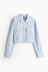 Camisa cropped celeste de algodón con cuello, botones ocultos, canesú con pliegue en la espalda, corte holgado, mangas largas, bolsillos superiores con solapa y botón, puños abotonados y bajo recto.