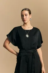 Top negro de manga corta con silueta amplia y caída fluida, confeccionado en un tejido liviano con leve brillo. Presenta un botón de nácar en la espalda con una pequeña abertura y mangas amplias tipo kimono.