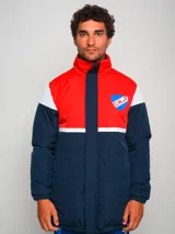 Campera parka de hombre con cuello alto, cierre frontal con cremallera y broches a presión, y escudo del Club Nacional de Football en el pecho. Diseño a bloques de color azul marino, blanco y rojo.