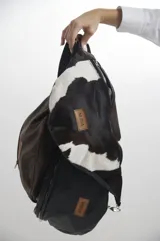 Riñonera de diseño combinado, con frente en cuero vacuno con pelo estampado tipo animal print de vaca en blanco y negro, y cuerpo principal en cuero liso color negro. Cuenta con cierre metálico, correa ajustable y etiqueta de cuero con logo grabado.