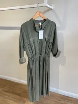 Vestido camisero largo color verde oliva, de pana fina con línea italiana. Tiene cuello mao, mangas largas con puños abotonados, cierre frontal con botones, dos bolsillos con solapa en el pecho, cintura ajustable con cordón y falda con vuelo.