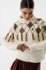 Sweater tejido de punto color crudo con cuello alto y diseño de rombos marrones con flecos.
