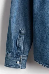 Camisa de denim azul de algodón rígido, corte holgado, cuello inglés, tapeta clásica con botones blancos, bolsillo de parche en el pecho, canesú en la espalda, mangas largas con puños abotonados y bajo levemente redondeado.