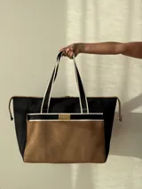 Bolso tipo tote de neopreno color negro y marrón, con cierre superior, bolsillo frontal marrón con logo rojo, bolsillo interno y externo con cierre, doble asa de mano marrón con detalles blancos y opción de correa larga.