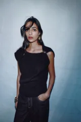 Jersey de punto negro, sin mangas (manga sisa), con cuello cuadrado drapeado y un broche dorado decorativo en el hombro.