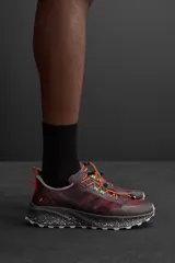 Zapatilla deportiva para carreras de montaña color borgoña con detalles en naranja y verde. Parte superior fabricada en tejido de punto transpirable con puntera y talón recubiertos de material Shield Protect Max. Ajuste de cordones mediante pieza técnica y suela con tecnología Spume Foam.
