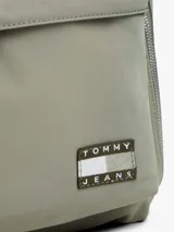 Mochila de nailon color verde oliva, con diseño liso y paneles texturizados. Presenta un compartimento principal con doble cremallera, un bolsillo frontal con cremallera y un parche con el logo de Tommy Jeans en la parte inferior. Las correas y el asa superior son negras.