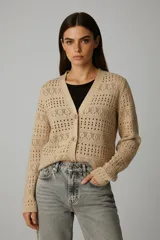 Cardigan de punto color beige, con escote en V y cierre frontal de botones. Presenta un tejido calado con patrones de agujeros y motivos en forma de 'X' y puntos.