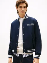 Campera estilo varsity azul marino con cuello, puños y ruedo acanalados con franjas blancas. Cierre frontal a presión y logo de Tommy Hilfiger bordado en blanco en el pecho.