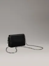 Cartera cruzada negra de piel sintética granulada, con correa de cadena plateada y cierre de solapa con placa metálica con el logo de Calvin Klein grabado.