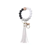Llavero con pulsera de cuentas redondas de silicona en colores blanco, gris y negro, con una cuenta de madera hexagonal y aro metálico dorado. Incluye borla de flecos de cuero sintético color blanco.