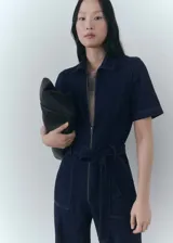 Mono largo de denim azul oscuro, con cuello camisero, mangas cortas y cierre frontal con cremallera. Incluye cinturón ajustable, bolsillos laterales y trabillas.