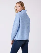 Buzo de punto grueso color beige, con cuello volcado y textura en relieve. Tiene mangas largas de corte hombro caído y puños anchos.