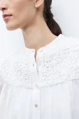 Camisa blanca de manga corta con cuello mao y detalles de bordado en el canesú superior. Presenta frunces en el pecho y la espalda, y un cierre frontal de botones.