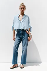 Camisa celeste y blanca a rayas verticales, confeccionada con lino e hilo metalizado. Tiene cuello solapa, mangas 3/4 con lazos en los puños y cierre frontal con botones ocultos por solapa.