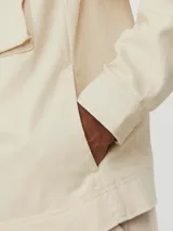 Chaqueta color crema, de manga larga, con cuello camisero y cierre frontal con cremallera metálica. Tiene dos bolsillos de parche con solapa en el pecho.