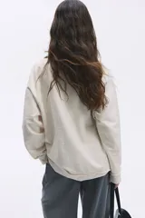 Buzo color crema de algodón con cuello camisero, tapeta de botones y mangas raglán. Tiene un pequeño bordado en el pecho y ribete desflecado en puños y bajo.