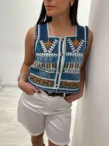 Chaleco corto de denim azul con bordado artesanal en tonos naranja, blanco y marrón. Tiene escote cuadrado, cierre de botones y terminaciones desflecadas.