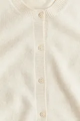 Cárdigan corto de punto fino color crema, con cuello redondo, tapeta de botones nacarados, manga corta y borde enrollado en puños y bajo.