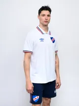 Remera polo blanca con cuello y puños con detalles tricolor (azul, blanco y rojo), cierre parcial con botones, logo de Umbro bordado en el pecho y escudo del Club Nacional de Football bordado en el pecho. Corte clásico.