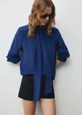 Blusa azul con lazo al cuello, de tejido fluido y satinado, con cierre oculto de botones y mangas largas abotonadas.