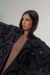 Ruana de tejido de punto grueso color gris oscuro con flecos en el borde inferior. Presenta apliques de lana en forma de líneas onduladas en color negro, fucsia, amarillo y celeste.