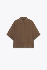Camisa color marrón de cuello solapa y manga corta con dobladillo. Tiene un corte holgado, bajo asimétrico con aberturas laterales y cierre frontal con botones ocultos.