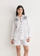 Camisa blanca de lino con bordados florales en los hombros, de manga larga y corte recto con abertura en los laterales. Cierre frontal con botones negros.