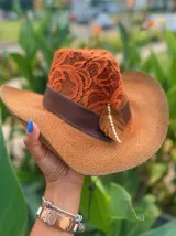 Sombrero de ala ancha tejido en hilo de coco y sintético, color marrón claro. La copa está cubierta con encaje naranja quemado con patrón floral. Tiene una cinta de cuero marrón oscuro y un aplique decorativo de hoja de gamuza con incrustaciones de pedrería.