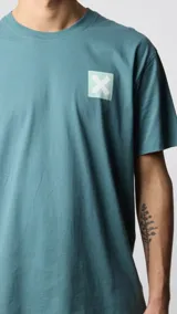 Camiseta celeste de algodón orgánico con cuello redondo y manga corta. Estampado en la espalda con un cuadrado turquesa y una cruz blanca en el centro.