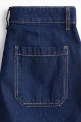 Shorts de denim azul oscuro, tiro alto, con cierre de botón dorado. Los bolsillos delanteros tienen un detalle de ribete trenzado.