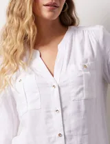Camisa blanca de lino y rayón, con cuello a la base y escote en V abotonado. Tiene dos bolsillos con botón en el pecho y mangas largas regulables con presilla y botón.