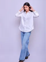 Camisa blanca de algodón, corte holgado, con cuello clásico y botones frontales. Las mangas son largas y se ven remangadas.